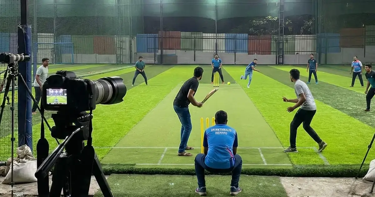 A GAMESWA camera on a tripod at the boundary of a floodlit turf ground, covering a cricket match in progress.